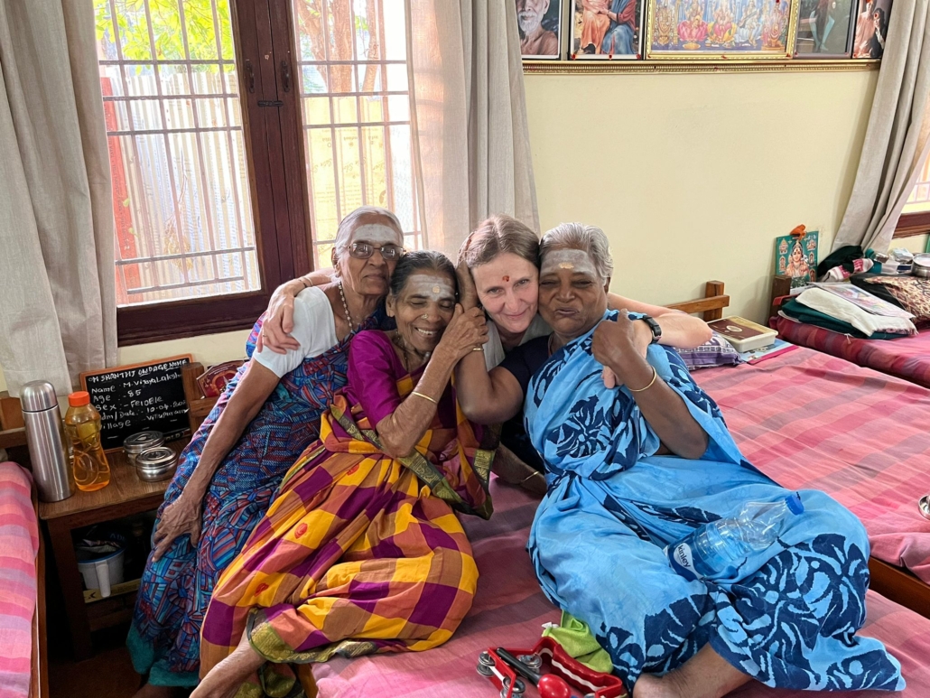 Four women embracing on a bed in an Indian care home, three elderly in sarees with forehead markings, one younger visitor