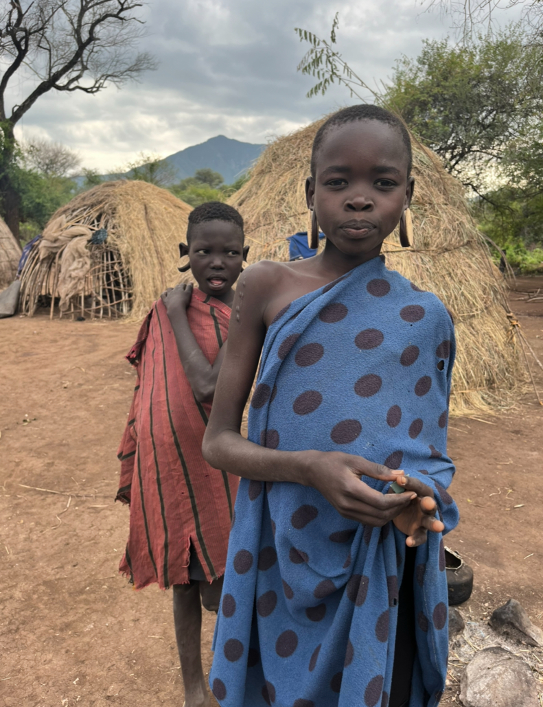 Two young African children in traditional cloth wraps standing in a village with thatched huts and mountains behind them
