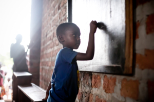 Young child in blue clothing writes on a chalkboard in a brick classroom with natural light