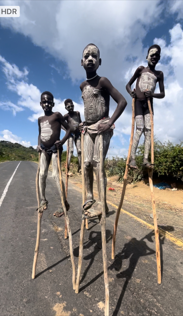Four children with traditional white body paint standing on wooden stilts on a paved road under a cloudy sky