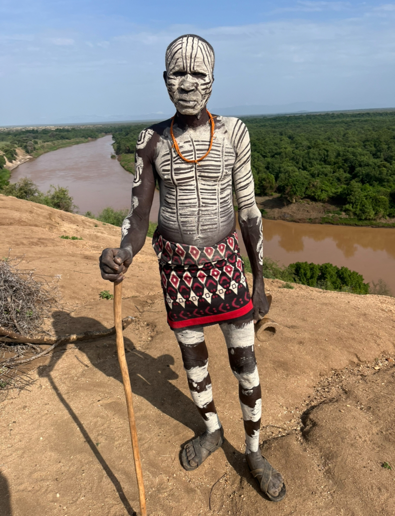 Man with traditional white body paint and patterned cloth wrap holds wooden staff on riverbank overlooking brown river and green forest