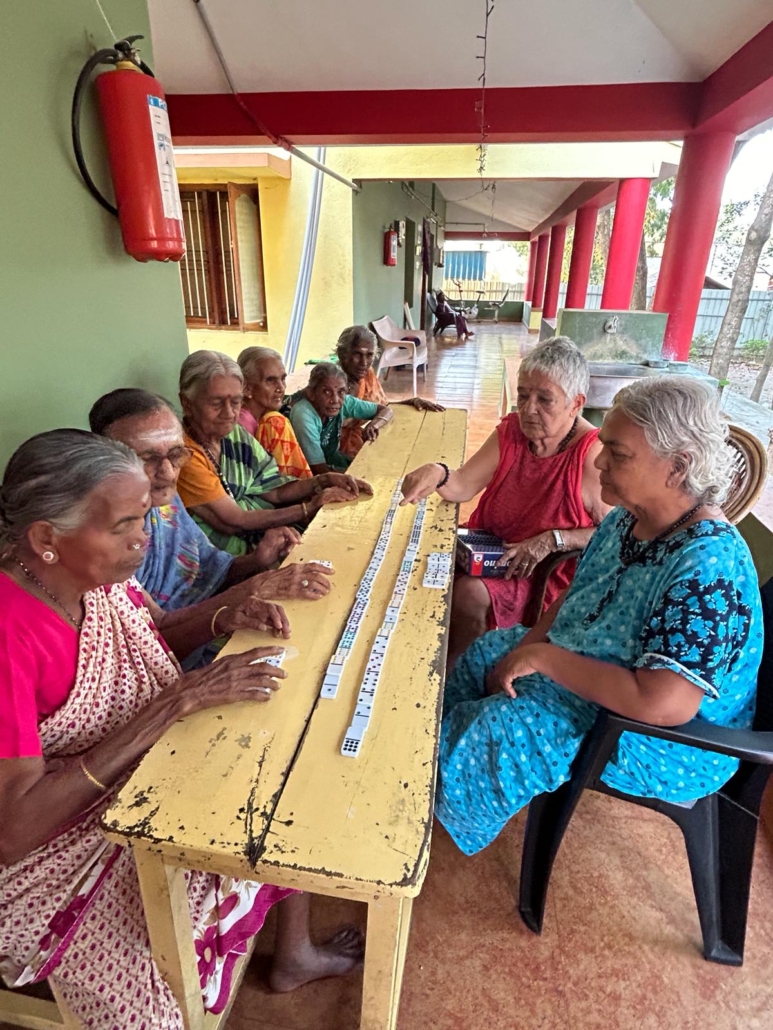 Elderly women sitting at a long yellow table playing dominoes together on a covered porch
