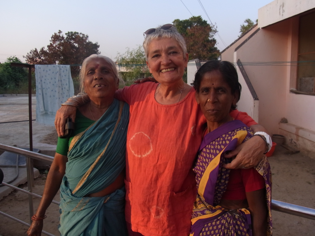 Three smiling older women with arms around each other, two wearing traditional Indian saris, one in orange tunic