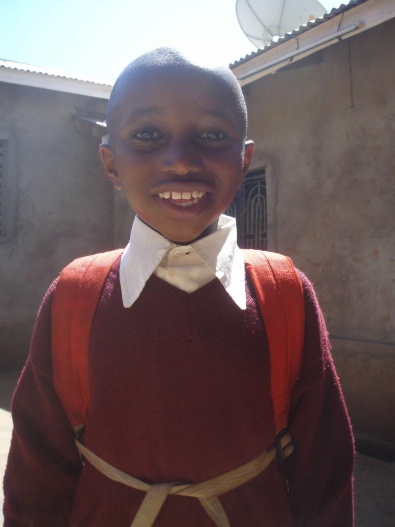 Smiling child in burgundy school sweater with white collar and orange backpack standing outside near building