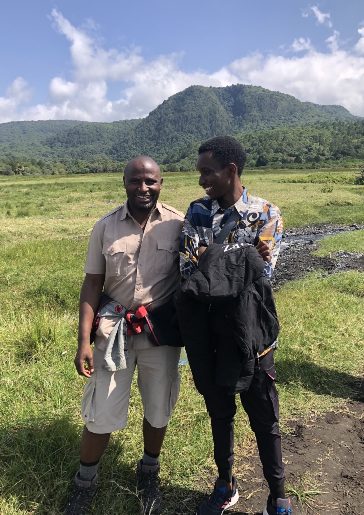 Two smiling men standing together in a grassy field with a lush green mountain in the background