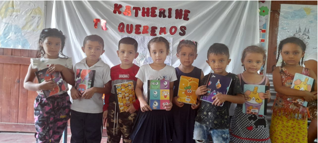 Eight children holding colorful notebooks stand in front of a banner reading Katherine Ti Queremos