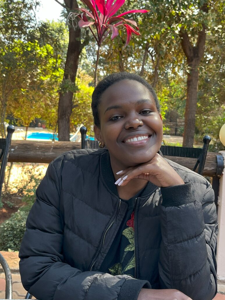 Young woman with short hair smiling, resting chin on hands, wearing black puffer jacket outdoors with trees and pool behind her