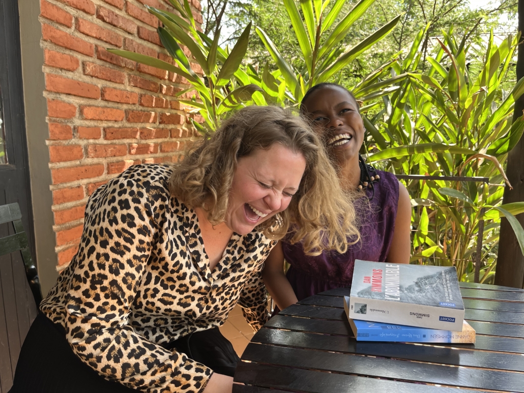 Two women laughing together at an outdoor table with books, one wearing leopard print, surrounded by tropical plants