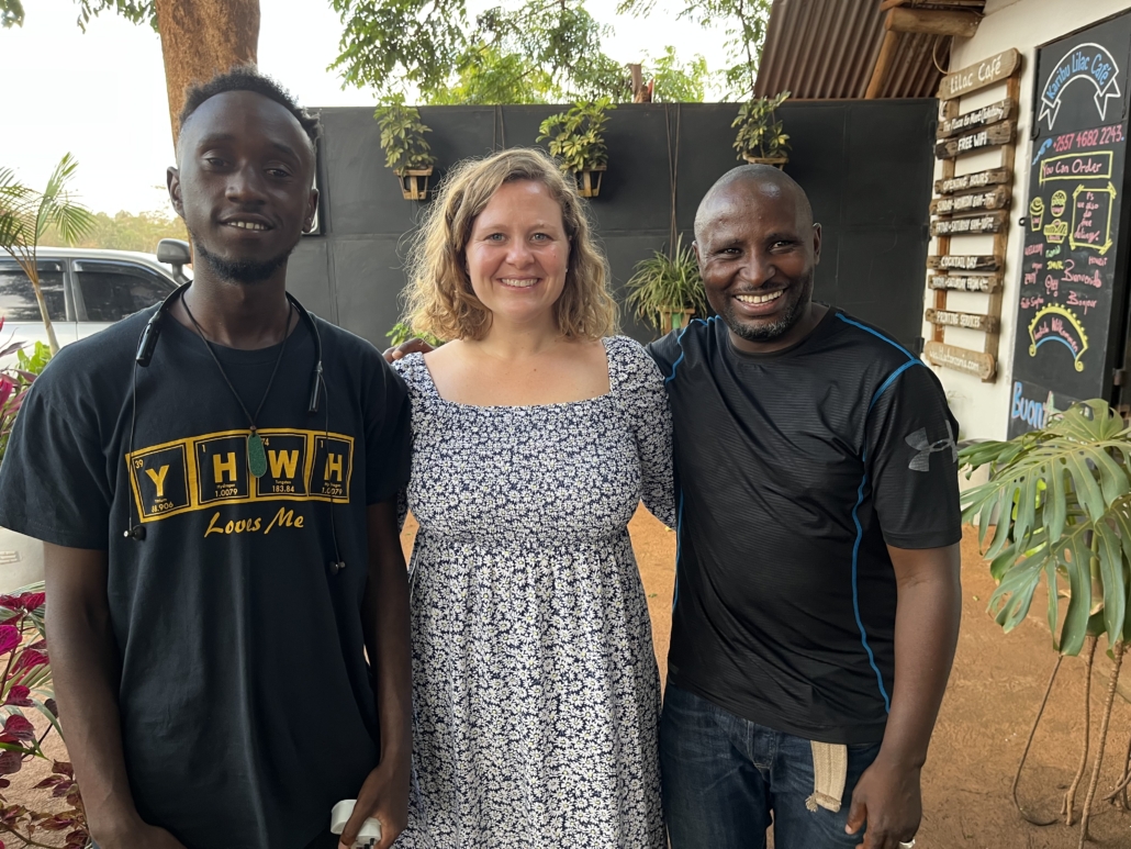 Three smiling people standing together at an outdoor cafe, two men in dark shirts and a woman in a floral dress