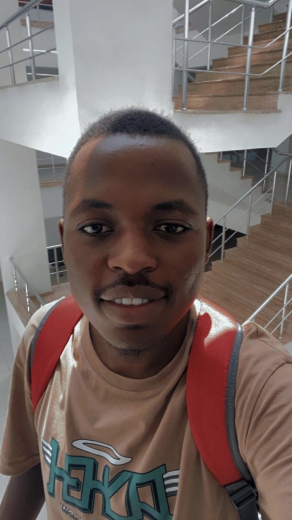 Young man taking selfie in modern building with wooden staircases, wearing tan graphic t-shirt and red backpack