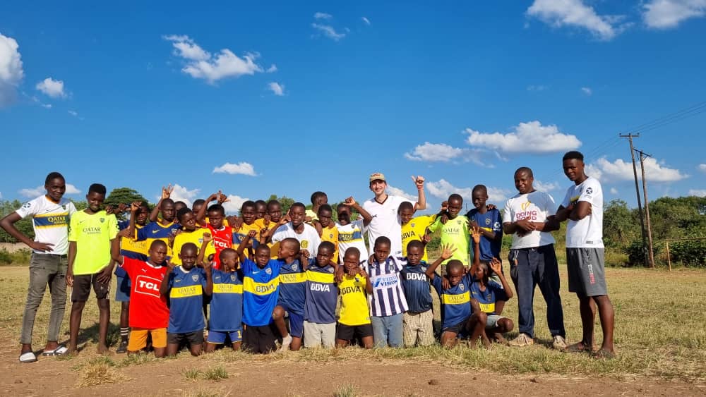 Group of children and adults in soccer jerseys posing together on a dusty field under blue sky