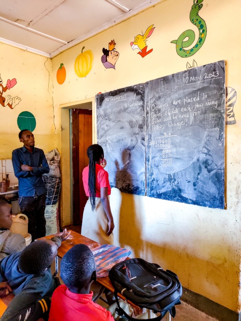 Student with braids works at chalkboard solving math division problem while teacher watches in decorated classroom