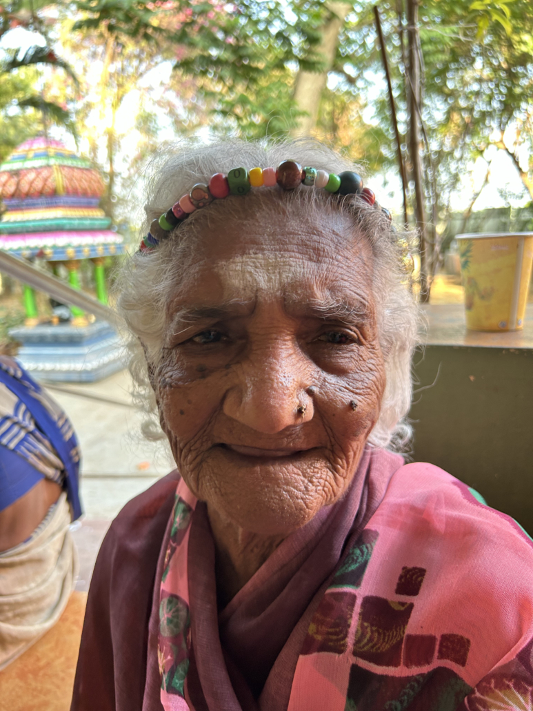 Elderly woman with white hair wearing colorful beaded headband, nose piercing, and pink floral sari, smiling gently