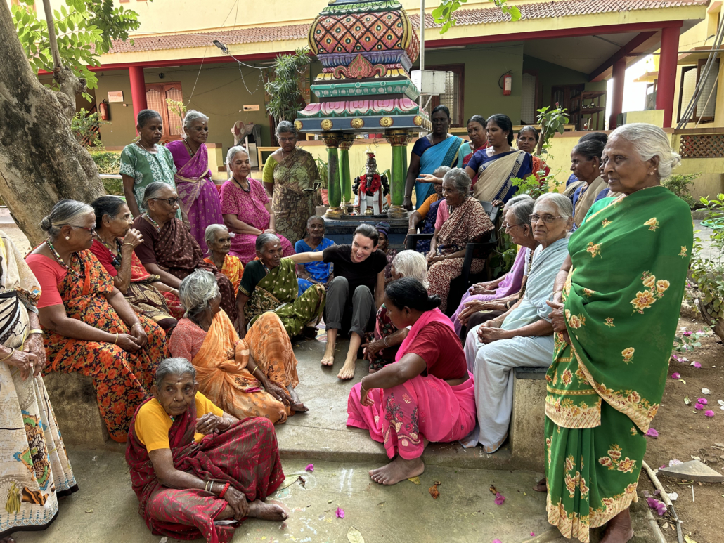 Group of elderly Indian women in colorful saris gathered around a small Hindu shrine with a Western visitor in black