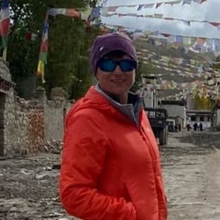 Person in orange jacket and purple beanie with blue sunglasses standing on mountain village path with colorful prayer flags