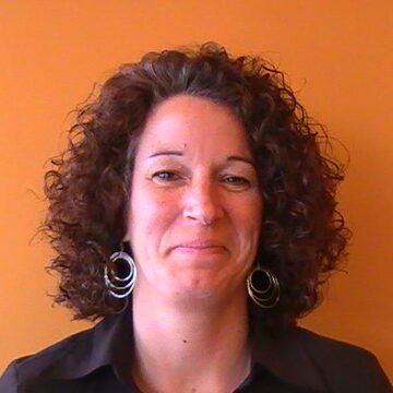 Woman with curly brown hair wearing a black shirt and silver hoop earrings, smiling against an orange background
