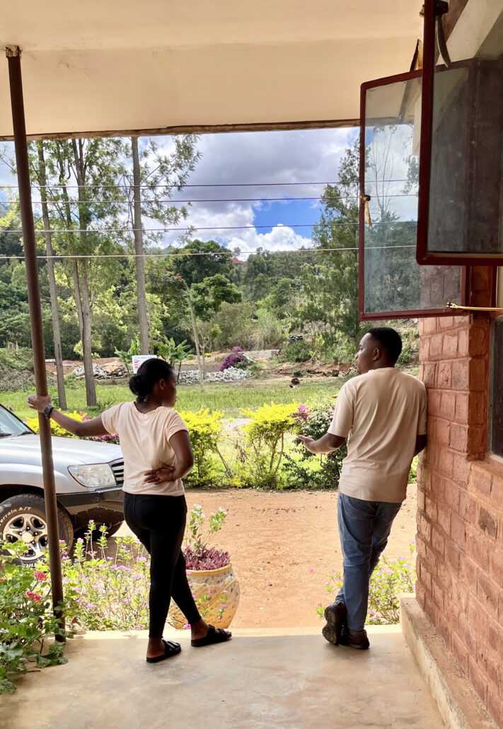 Two people in light shirts stand on a covered porch looking out at a green landscape with trees under a cloudy blue sky