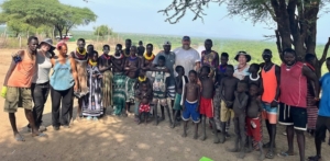 Group of tourists and indigenous villagers posing together under a tree in an African savanna landscape