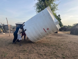 Three people push a large white water storage tank upright in a rural village with thatched huts