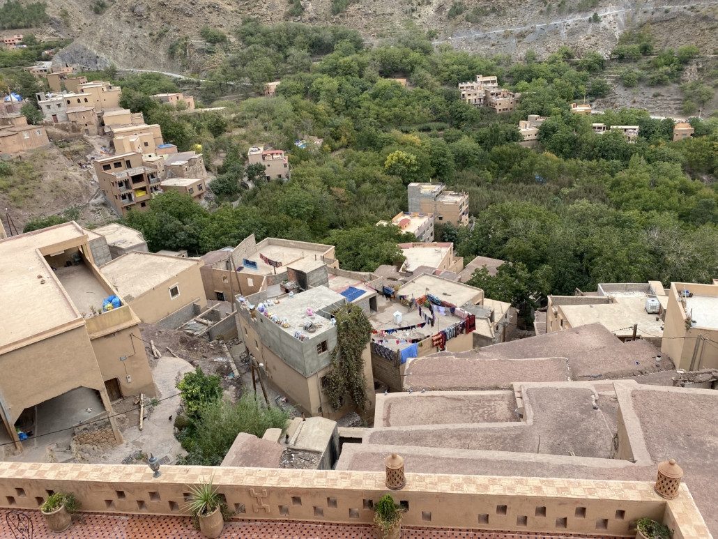 Aerial view of a hillside village with tan adobe buildings, flat roofs with laundry drying, and lush green trees