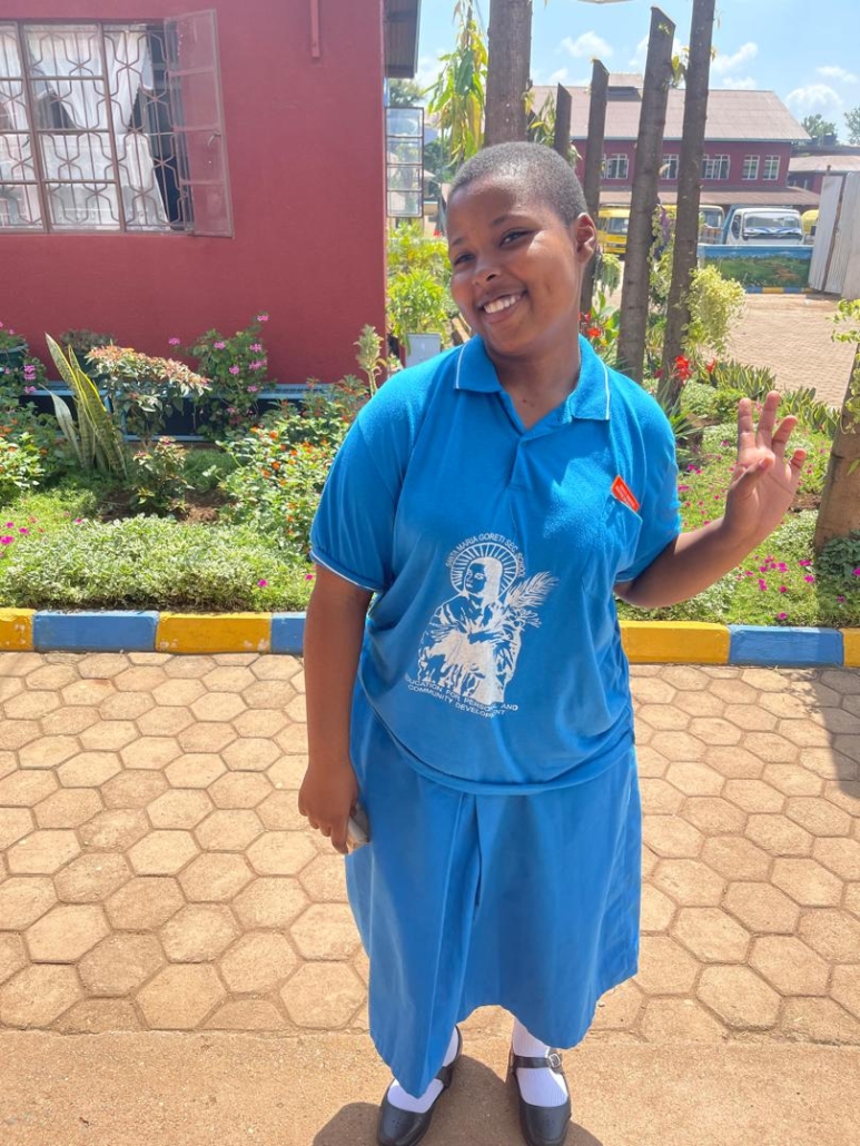 Smiling student in blue school uniform making peace sign, standing on paved walkway with gardens and buildings behind