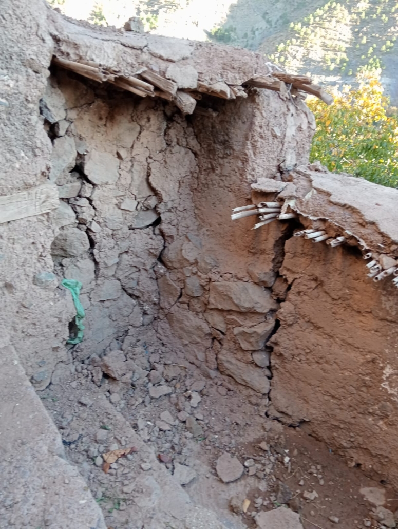 Collapsed mud and stone wall of traditional building with exposed wooden beams, rubble, and mountainous terrain in background