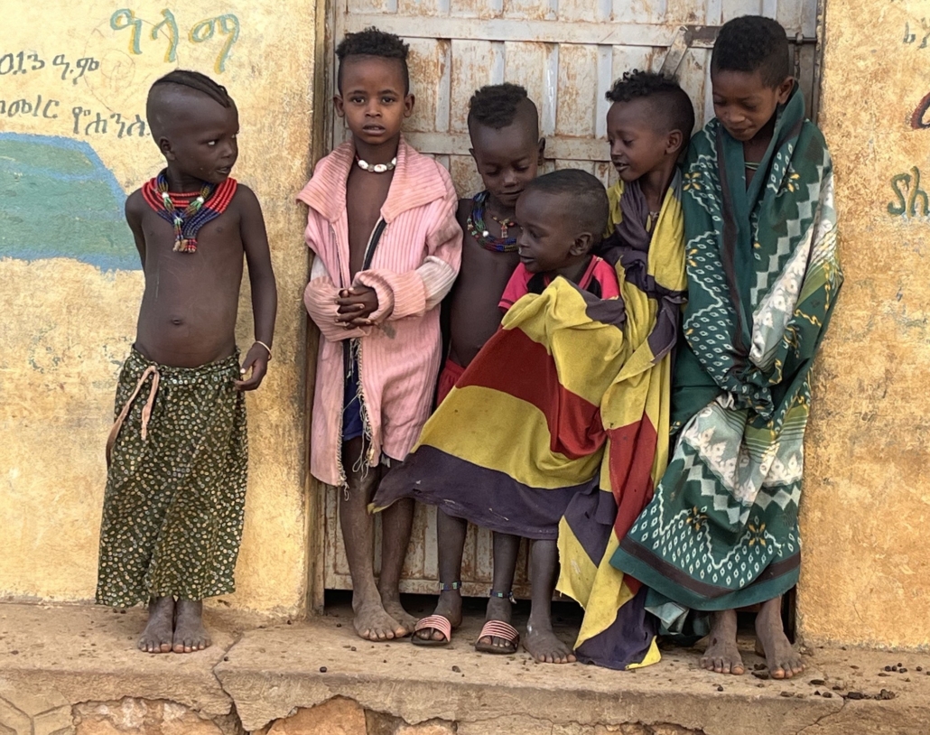 Six African children standing barefoot by a yellow building doorway, wearing colorful traditional clothing and beaded necklaces