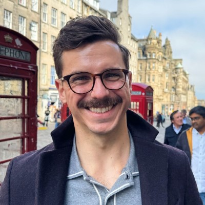 Smiling man with glasses and mustache wearing gray sweater and dark coat on Edinburgh street with red phone booth