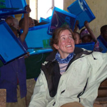 Woman in light jacket smiling while children behind her hold up blue plastic boxes above their heads