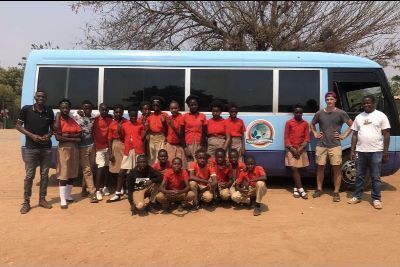 School group in red uniforms poses in front of blue bus with logo on dusty ground with trees in background