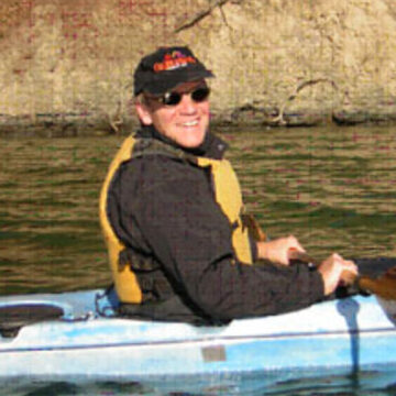 Person in sunglasses and yellow life vest smiling while sitting in a blue kayak on calm water