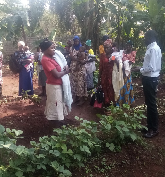 Group of women with babies gathered outdoors near a vegetable garden, listening to a man in white shirt speaking